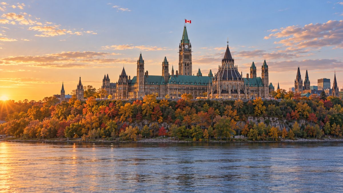 Parliament Hill in Ottawa at sunset, seen across the Ottawa River, with autumn trees along the cliff.